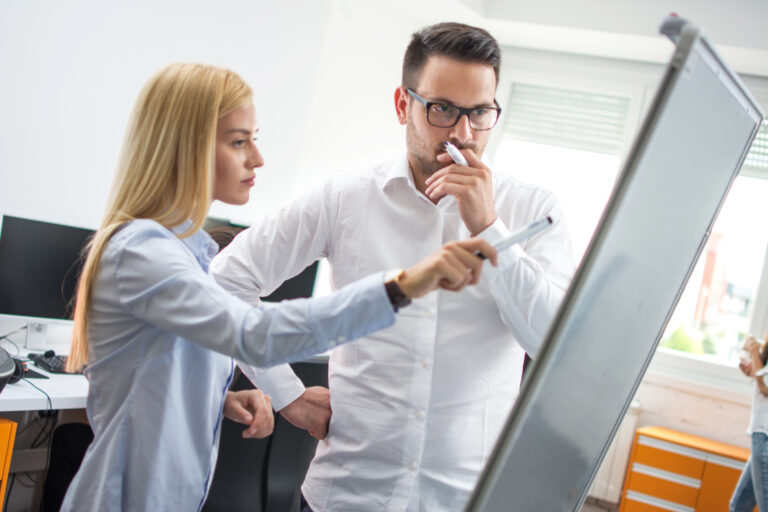 Businesswoman pointing with marker to flipboard and explaining company’s financial situation to male pensive colleague ACH vs Echeck